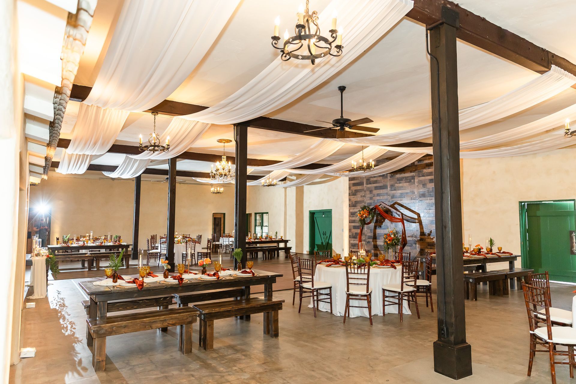 Spacious banquet hall with draped white ceiling, chandeliers, wooden tables, and green doors