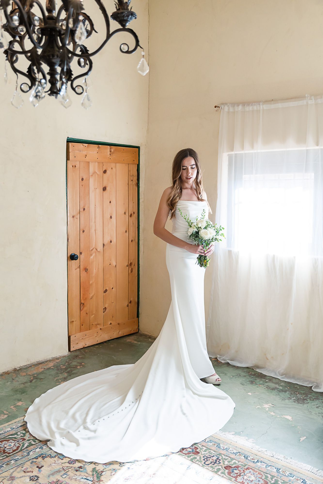 Bride in a white gown holding a bouquet in a bright room by a window and wooden door