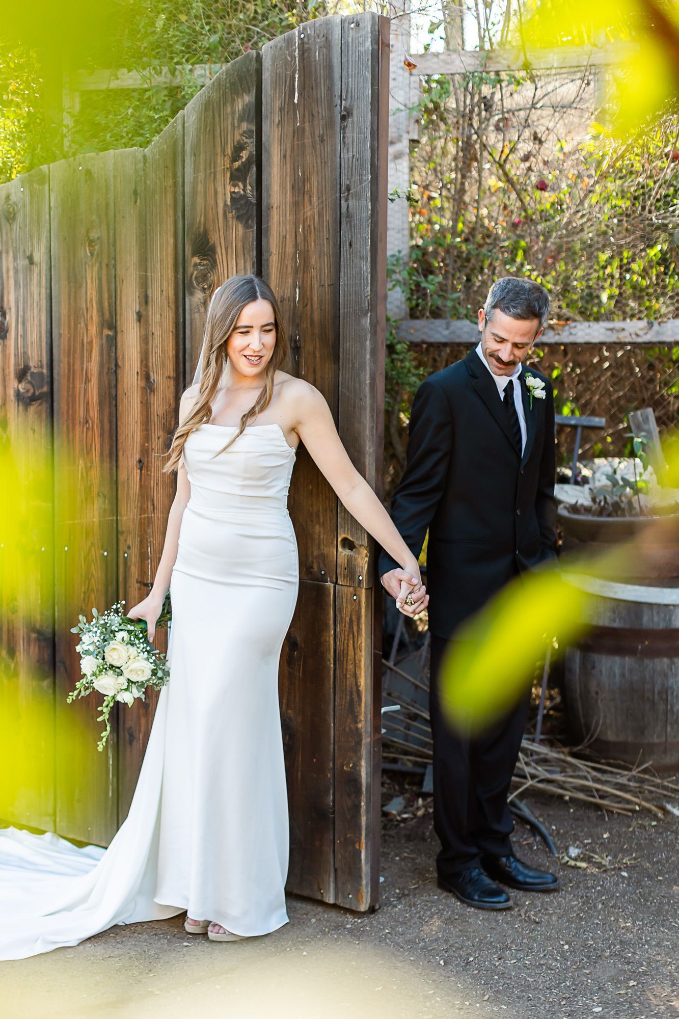 Bride in white dress and groom in black suit holding hands by a wooden gate outdoors