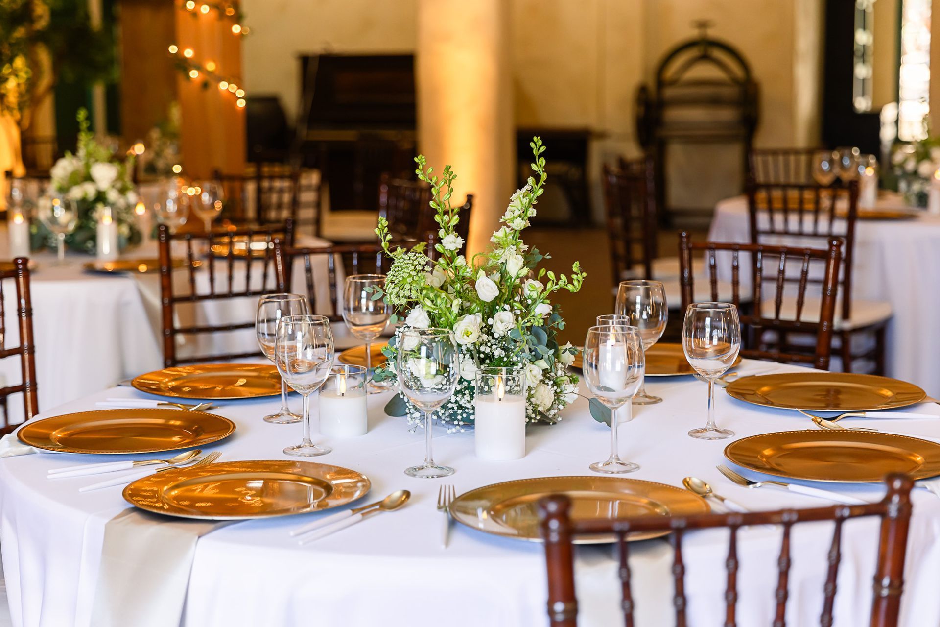 Elegant banquet table with white tablecloth, gold plates, glassware, and floral centerpiece in a warm-lit hall