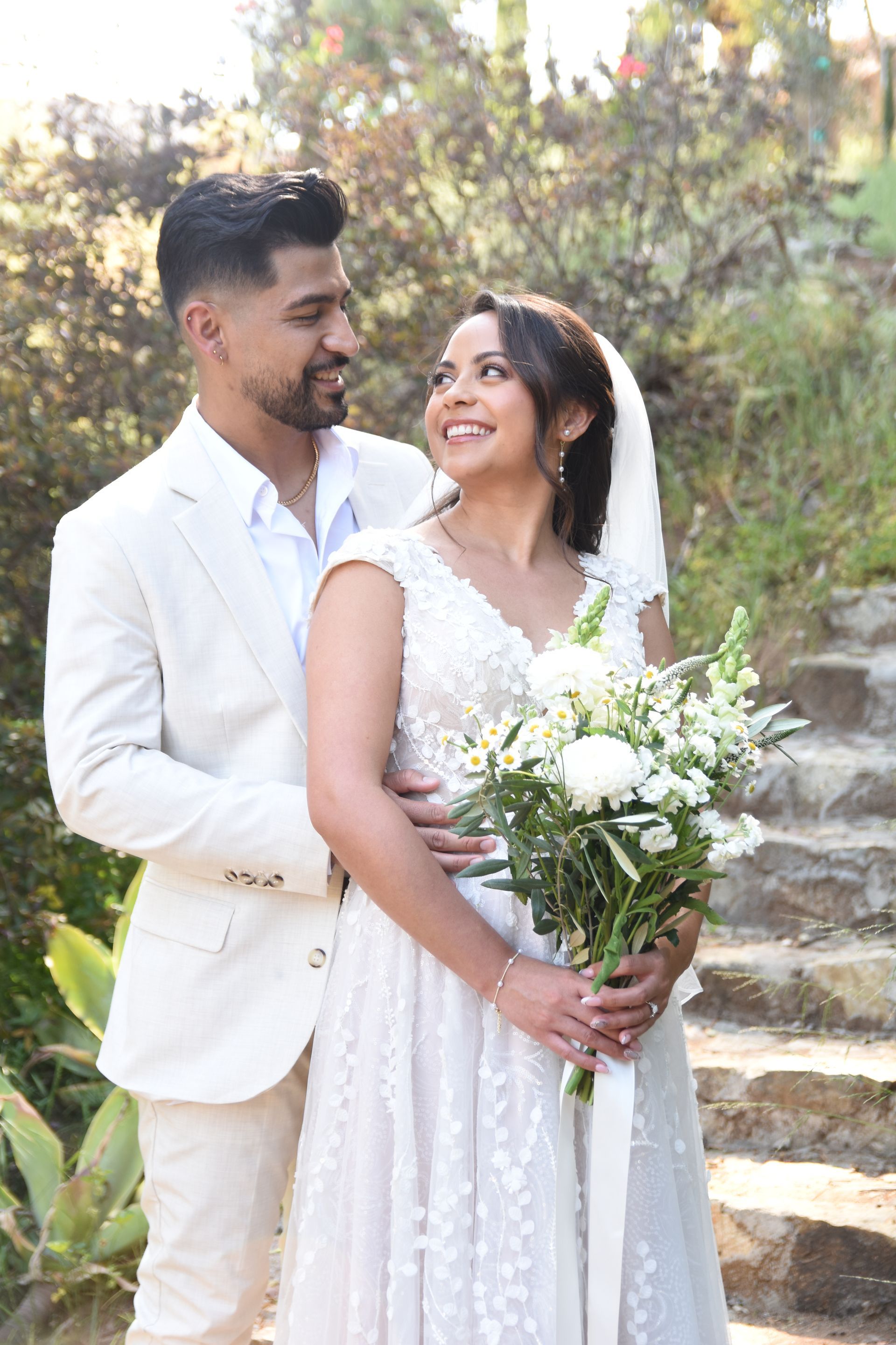 Bride and groom in white outfits smiling outdoors, holding a bouquet on stone steps