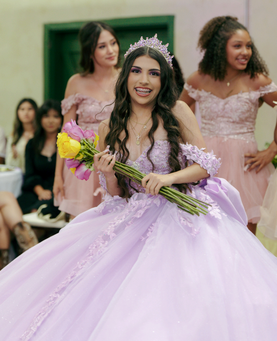 Young woman in a lavender gown holding flowers, with two women in pink dresses behind her at an indoor celebration.