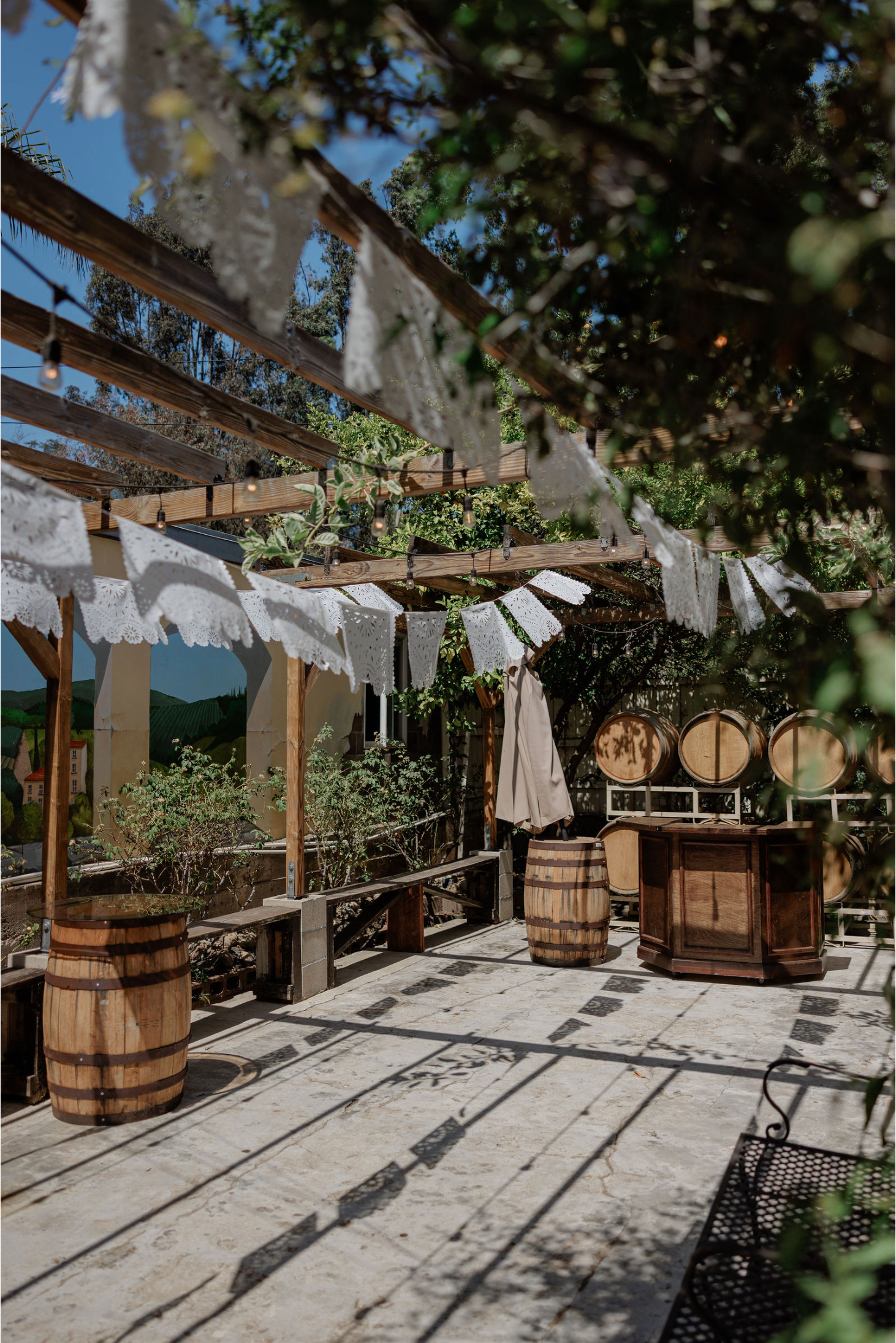 Outdoor rustic reception area with wooden barrels, white bunting, and string lights under trees