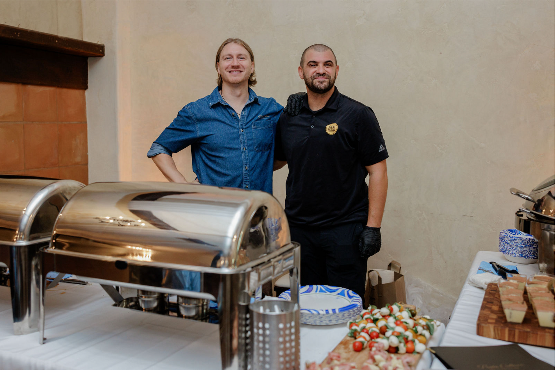 Two men standing behind a buffet table with chafing dishes and food trays at an indoor event