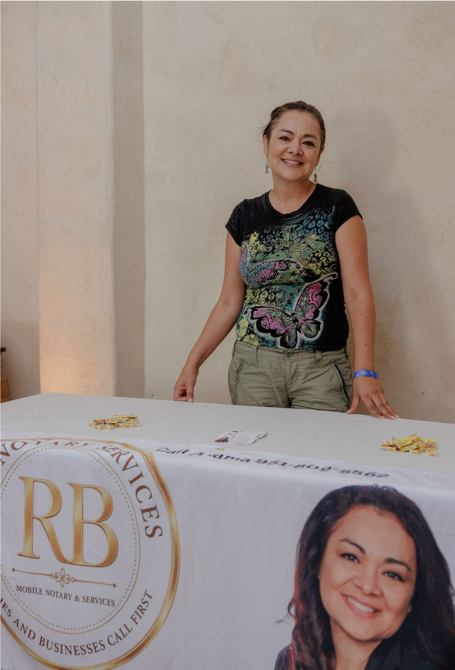 Woman standing behind a table with a banner and portrait, smiling in a beige indoor setting