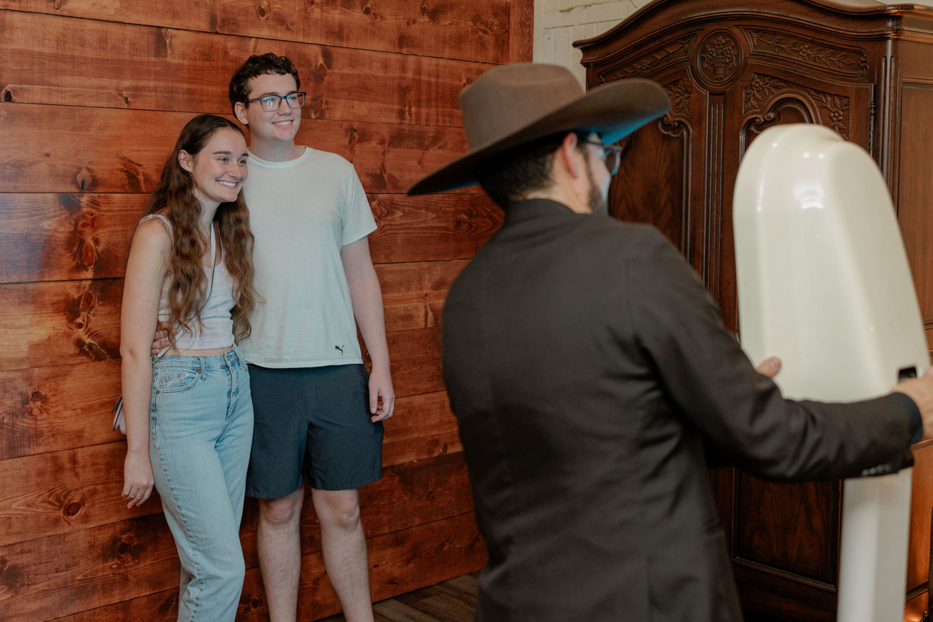 Couple posing by a wooden wardrobe while a person in a hat adjusts a white dress form