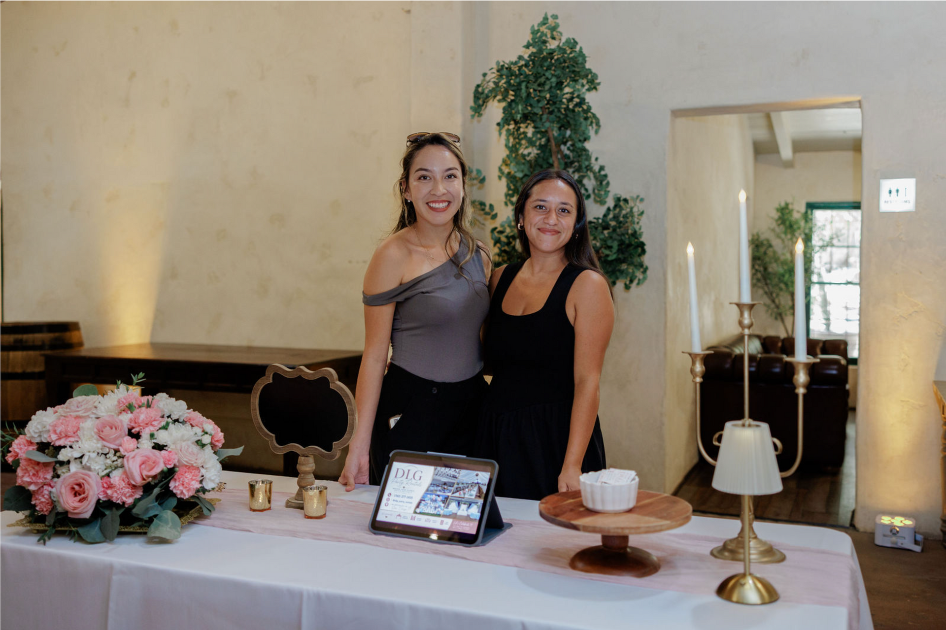 Two women standing behind a display table with flowers, cakes, and decor in a bright room