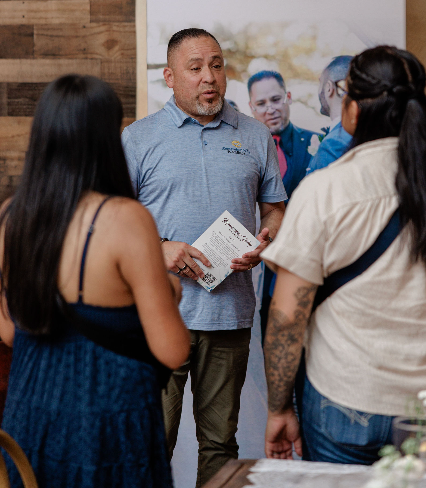 Man speaking to two people at an indoor event, holding brochures and facing a booth display.