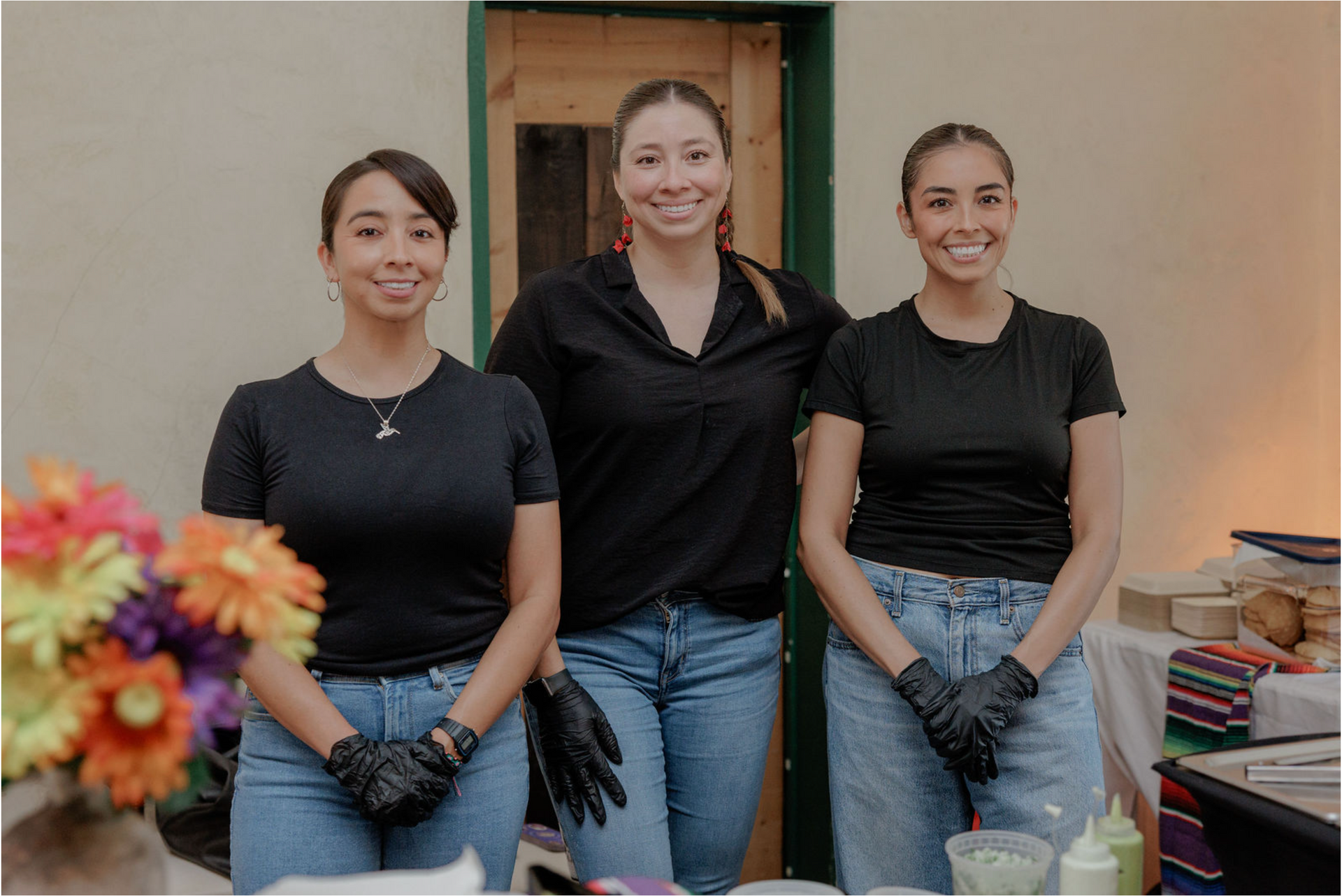 Three smiling volunteers in black shirts and jeans stand behind a table with flowers and supplies.
