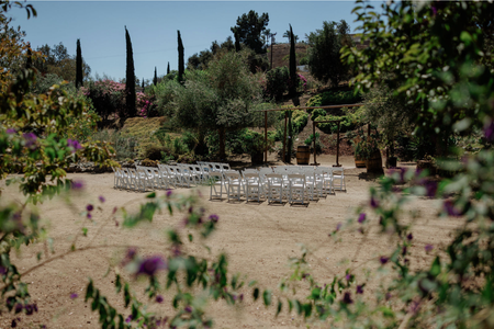 Outdoor garden ceremony setup with white chairs arranged on a sandy area surrounded by trees and flowers