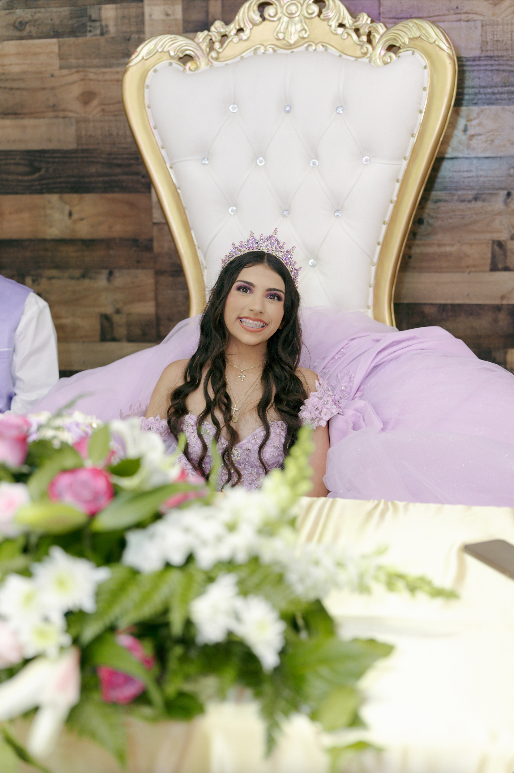 Woman in a lavender dress seated on a white tufted chair, with flowers blurred in the foreground.