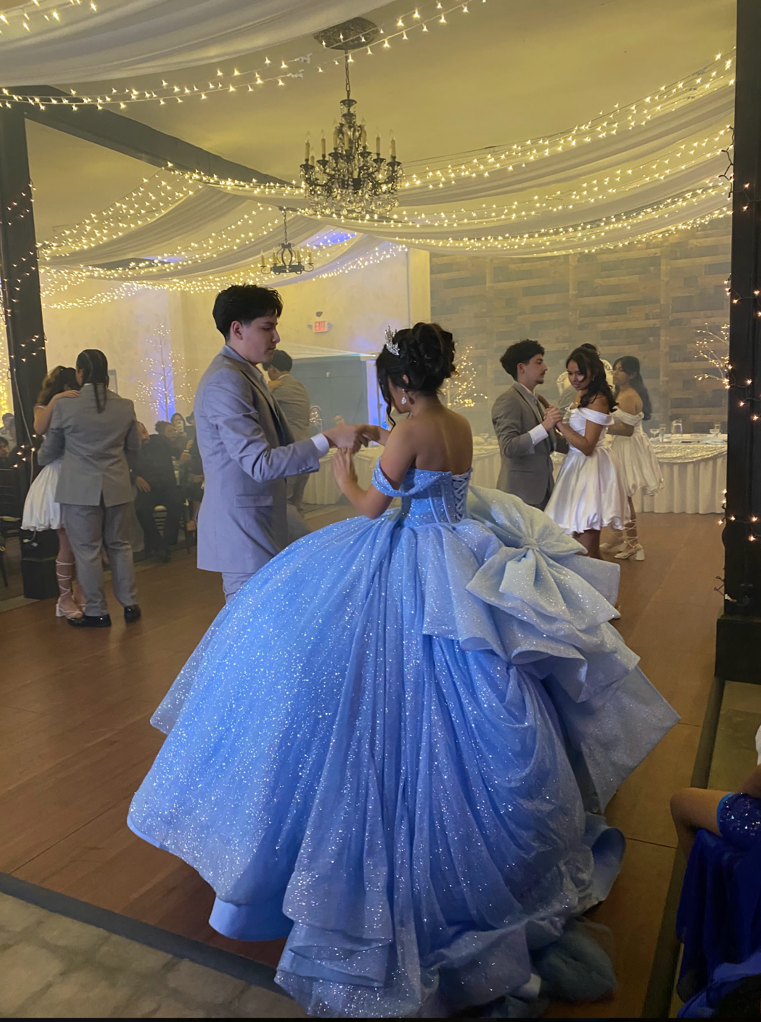 Couple dancing at a formal ballroom party, the woman in a sparkling blue gown under warm string lights.