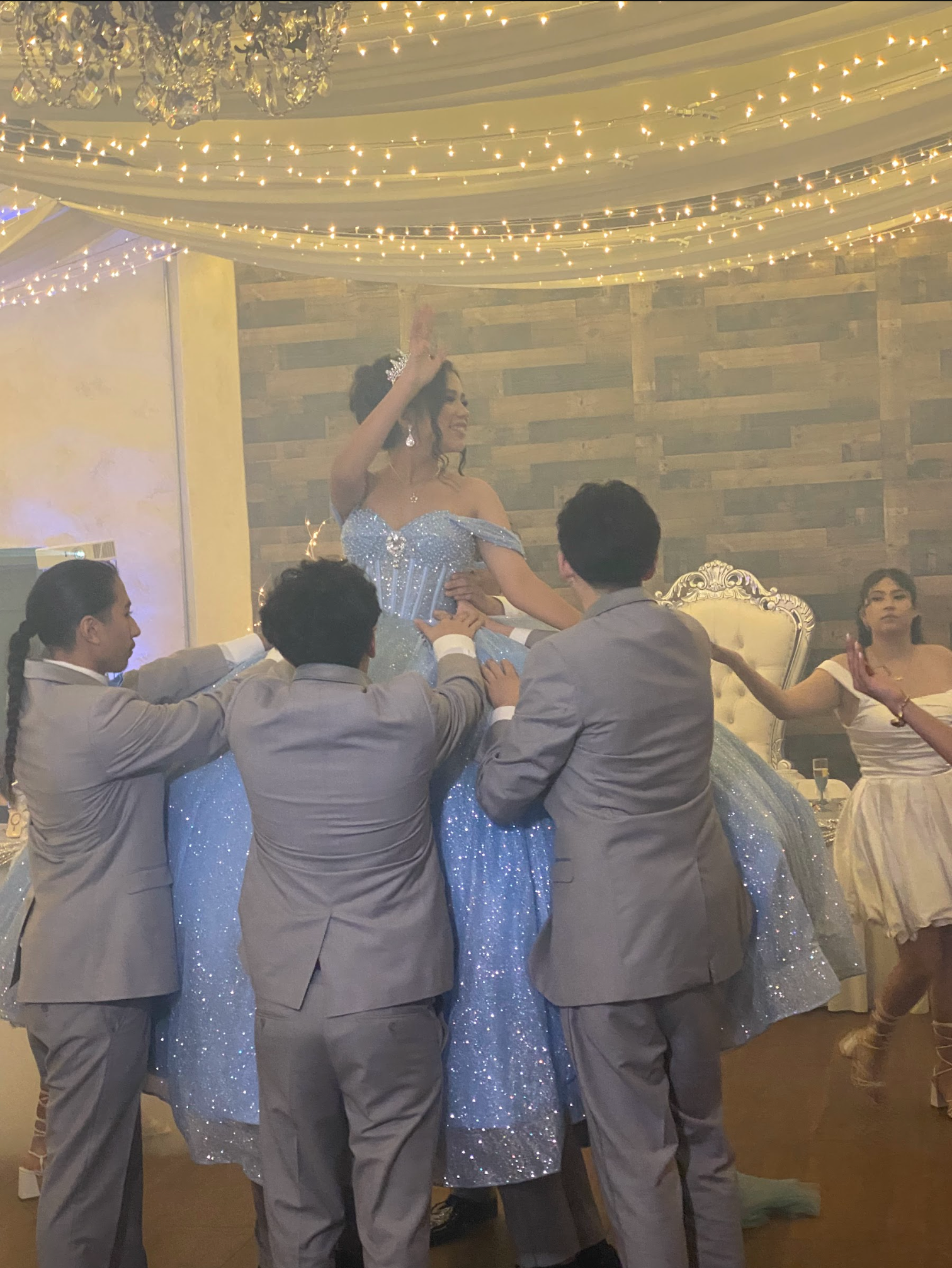 People in formal attire lift a sparkling blue-dressed dancer at a decorated indoor celebration.