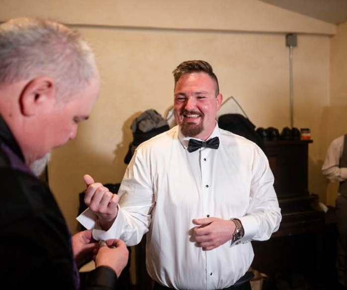 Smiling man in white tuxedo shirt and black bow tie holding a small item at an indoor event
