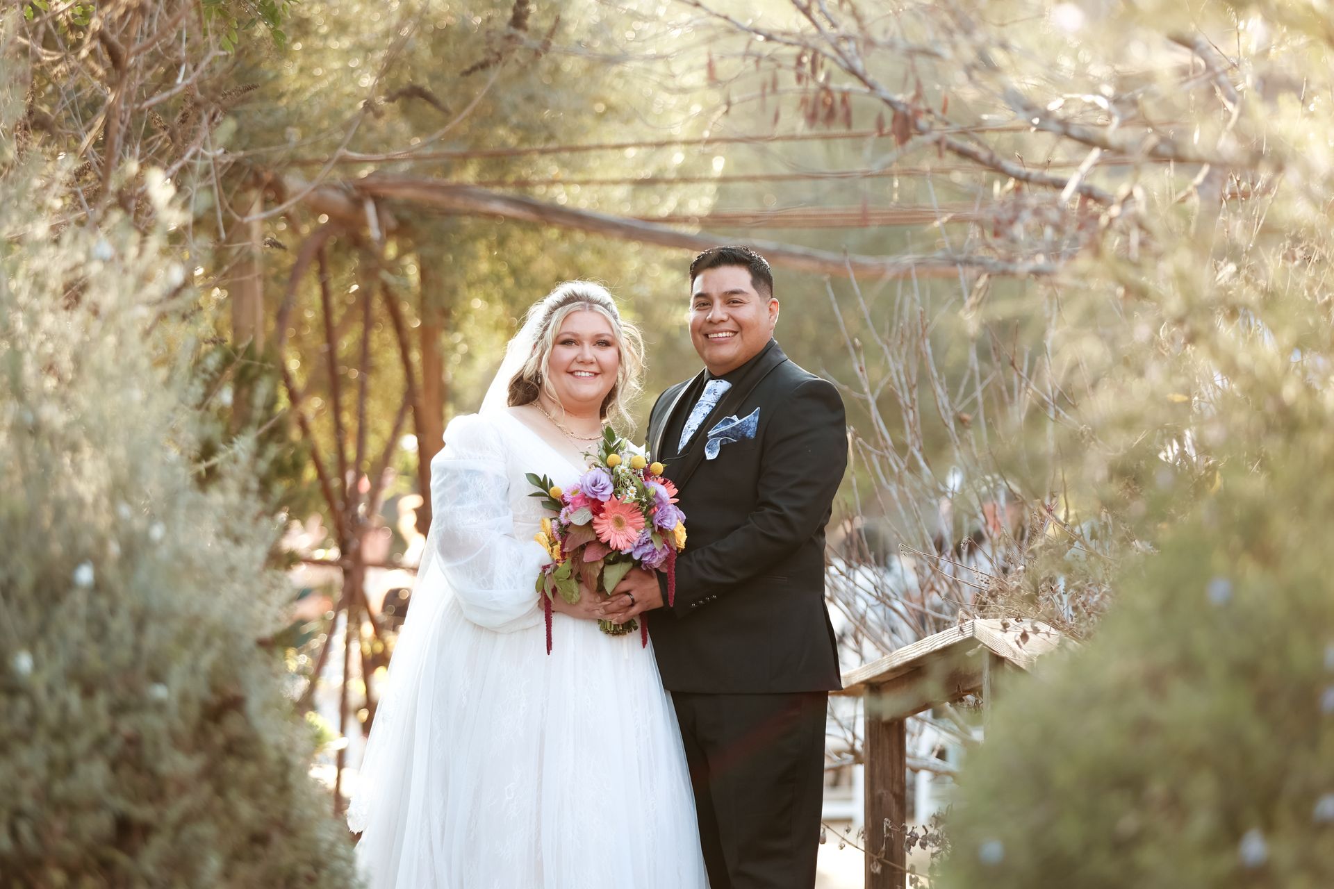 Bride and groom pose with a bouquet in a wooded outdoor setting on a wooden bridge.