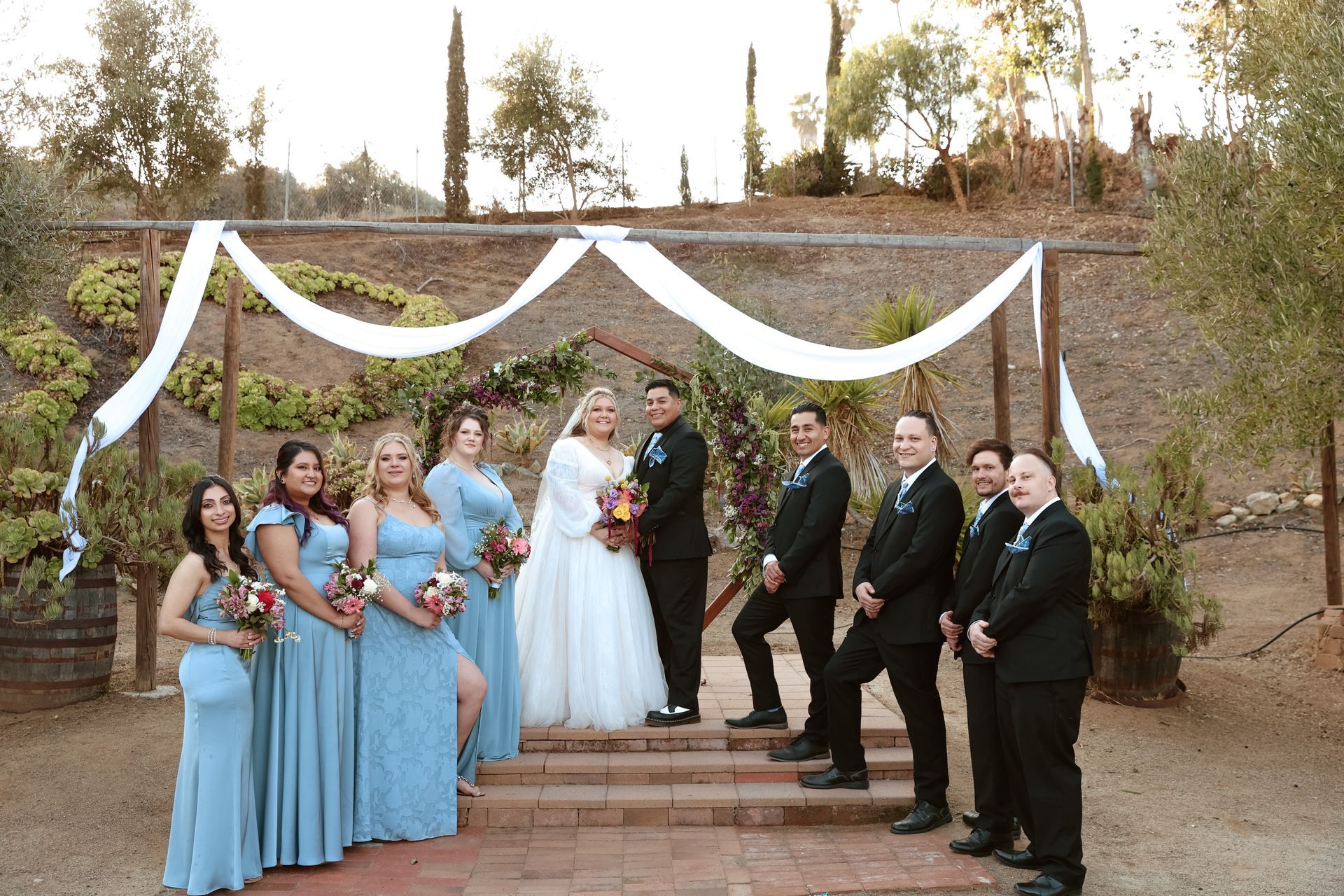 Wedding party posing outdoors under white drapery, with bridesmaids in blue and groomsmen in black suits.