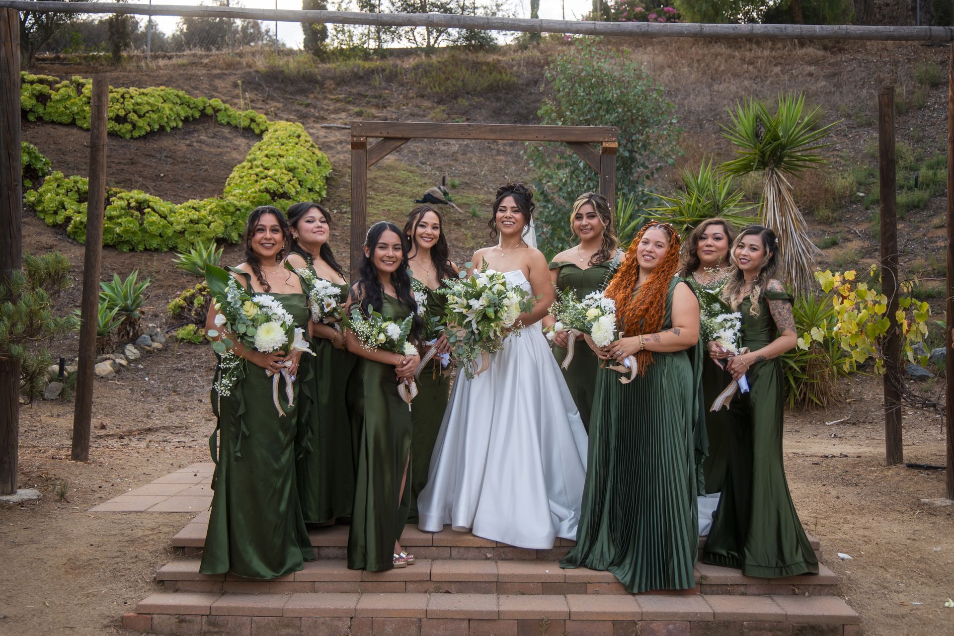 Wedding party posing outdoors in dark green dresses with the bride in a white gown, holding bouquets.