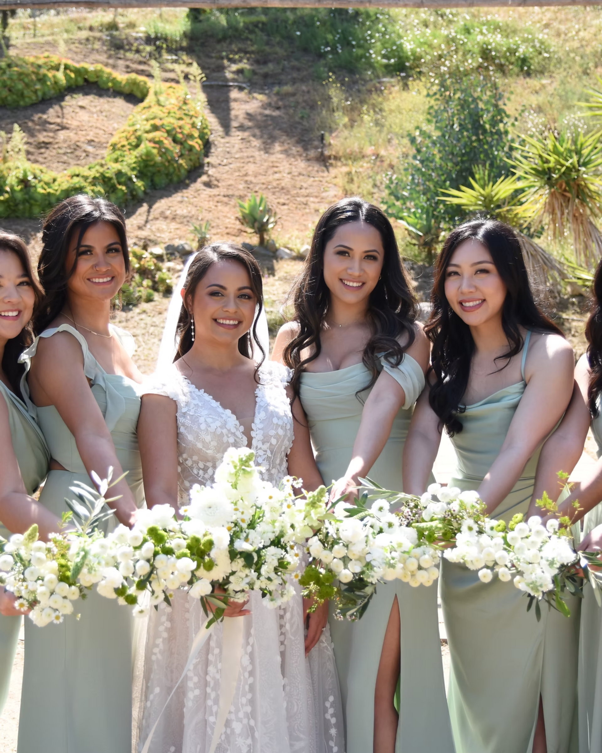 Five women in pastel dresses hold white floral bouquets outdoors at a sunny wedding ceremony.