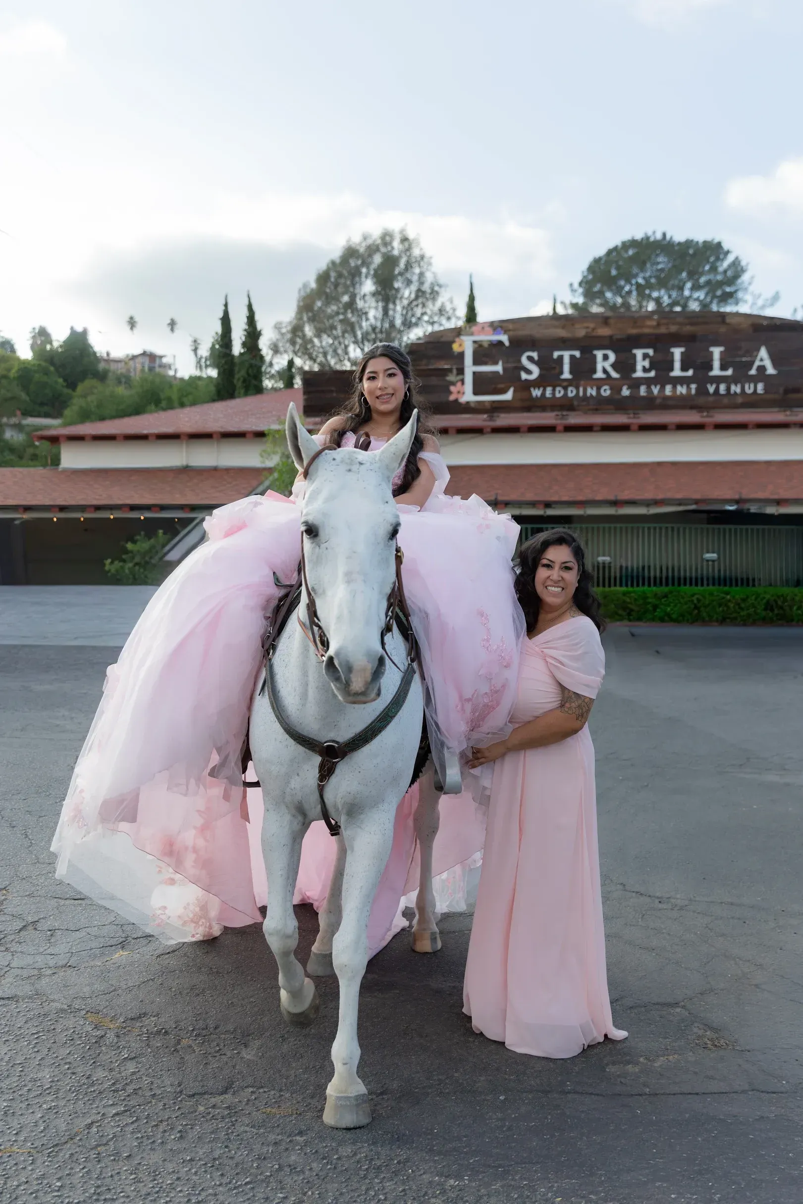Woman in a pink gown on a white horse with a pink-dressed companion, in front of an Estella sign.