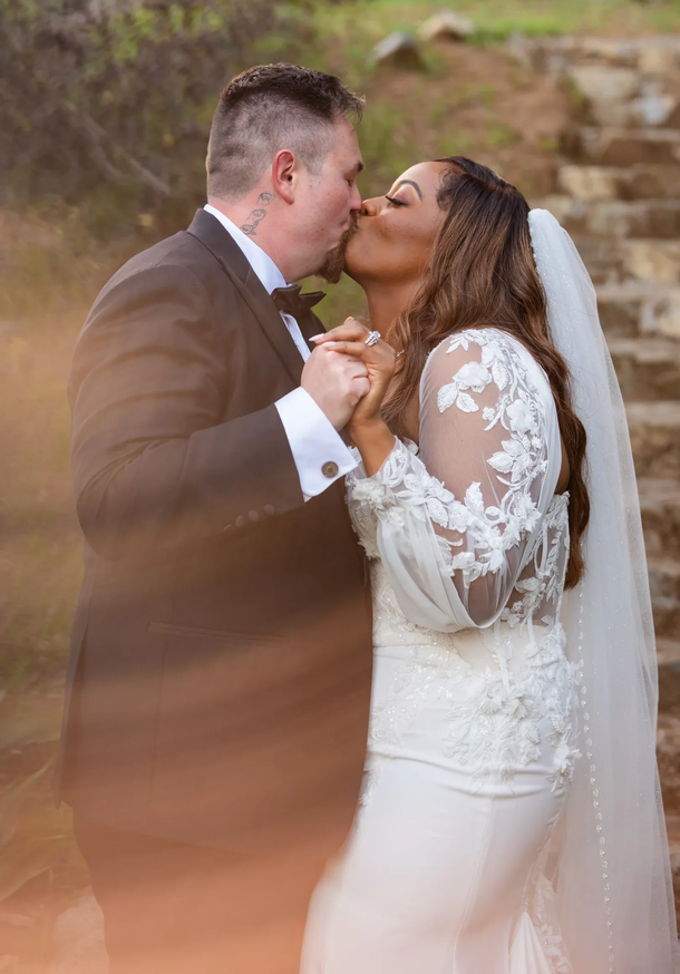 Couple in wedding attire kissing and holding hands outdoors, with a soft blurred foreground.