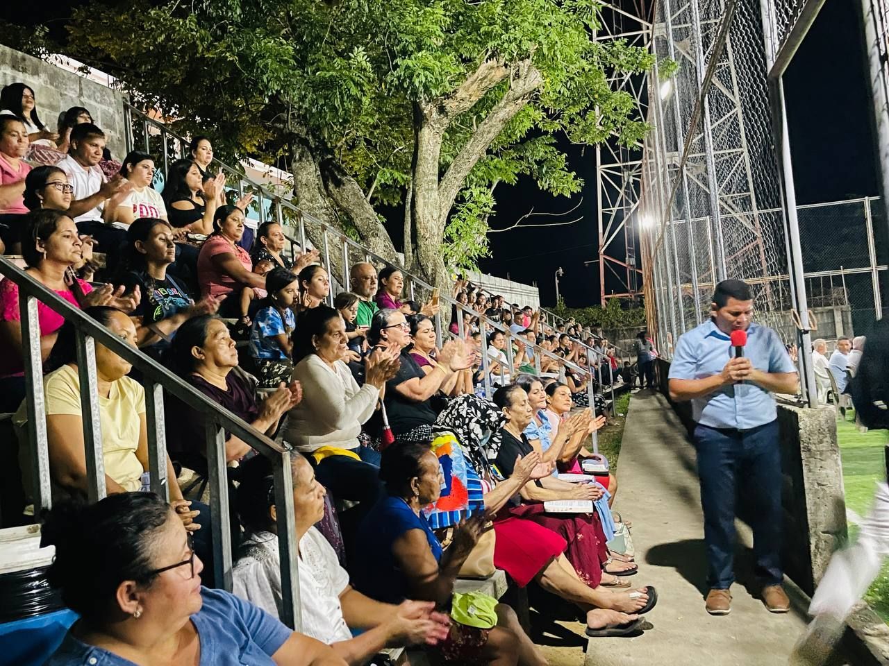 Crowd seated outdoors at night, watching a man in a blue shirt speak into a microphone near a stage