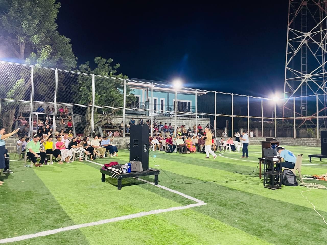 Outdoor nighttime gathering on a lit sports field, with many people seated near a chain-link fence and bleachers.