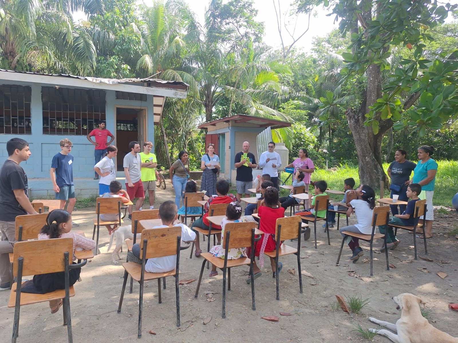Outdoor classroom with children and adults gathered around desks under trees near a small building