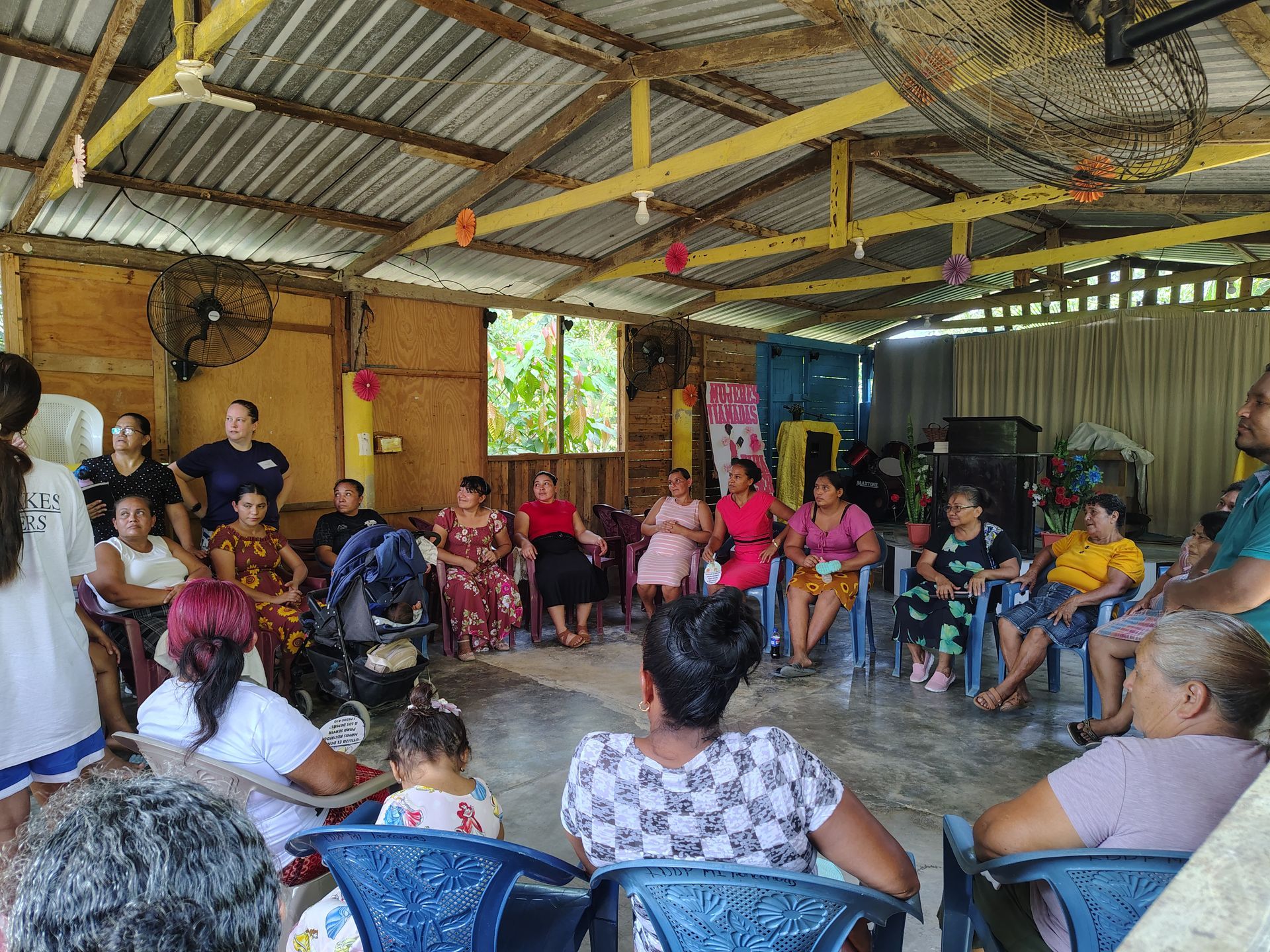People seated in a rustic hall during a community meeting, with a speaker at the front.
