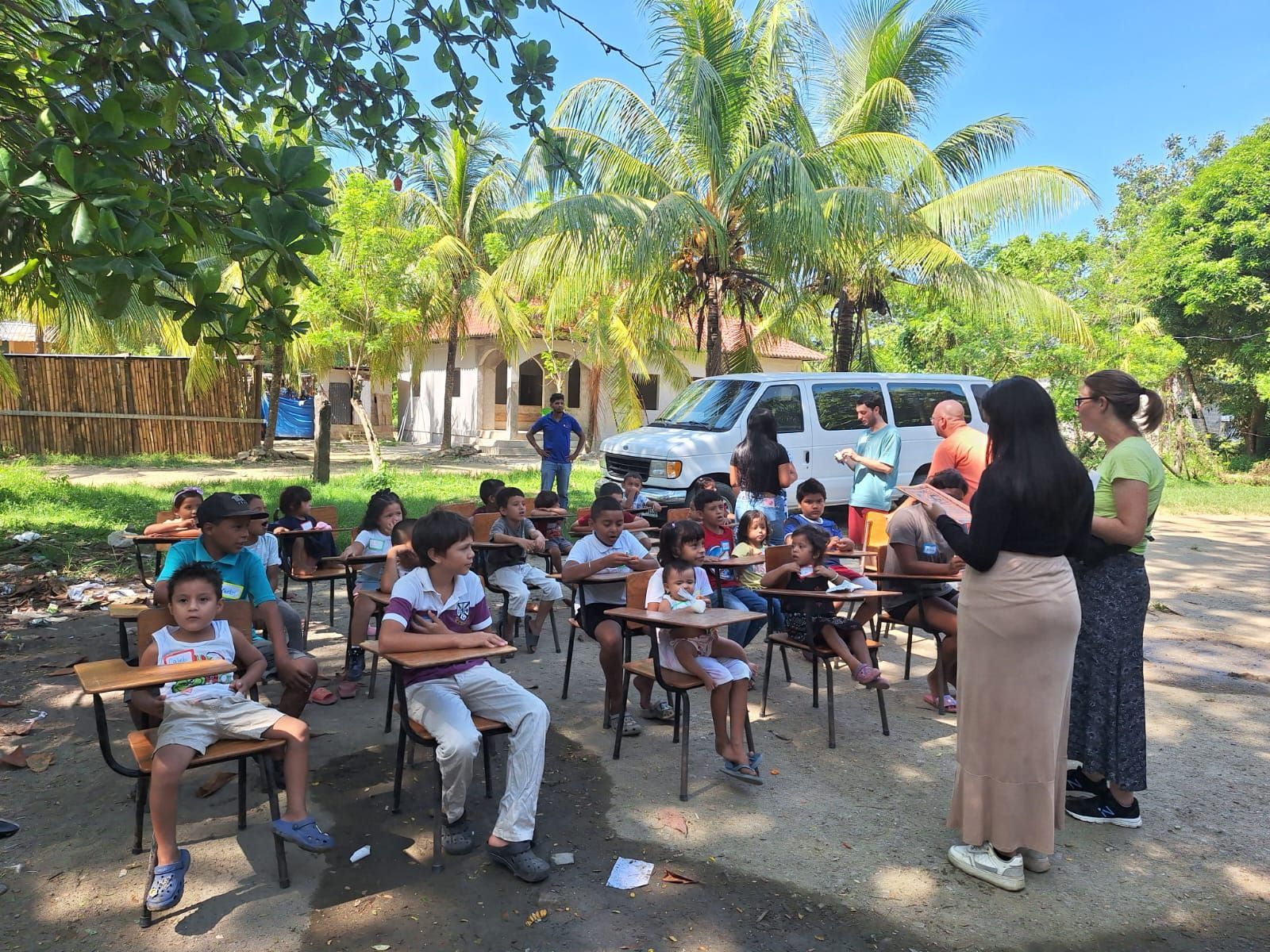 Teacher speaking to children seated at desks outdoors under trees, with a van and school building nearby.