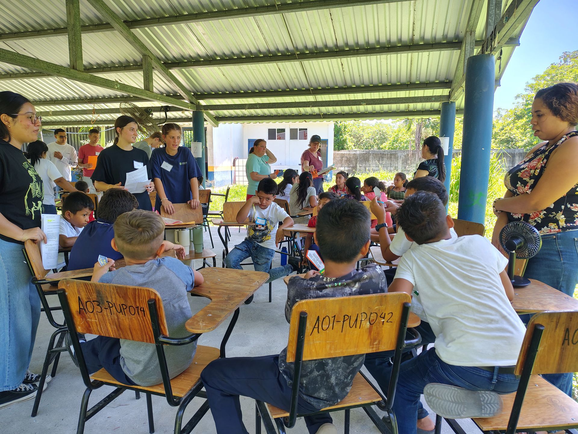 People seated around tables in a covered outdoor meeting space, with presenters standing at the front.