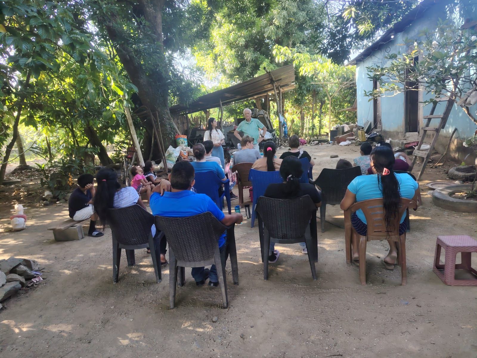 People seated outdoors on plastic chairs under trees, listening at a small gathering.