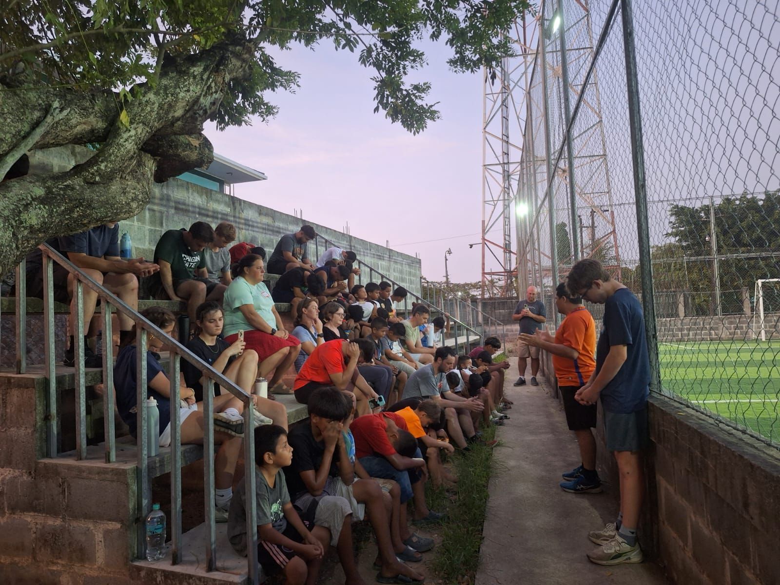 Crowd seated on bleachers beside a sports field, watching a coach speak near the fence.