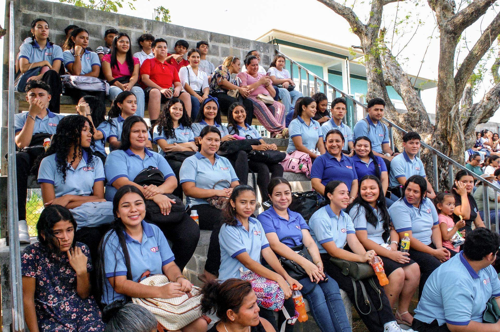 Crowd of students in blue uniforms seated on outdoor steps, listening during a school gathering