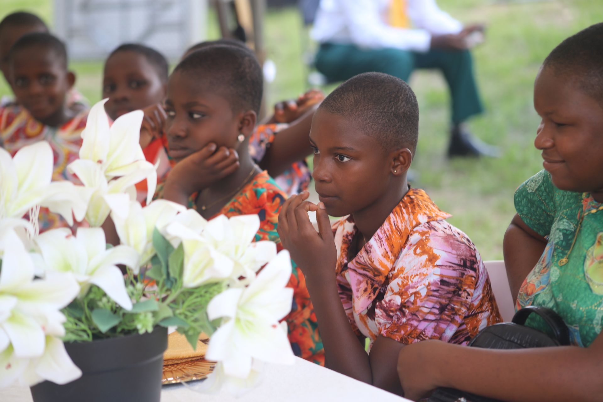 Children seated at an outdoor table, looking at white lilies in a black vase at a gathering