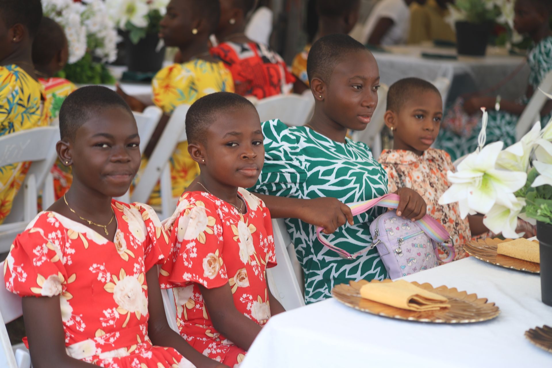 Children seated at a decorated table at an event, with flowers and plates in the foreground.
