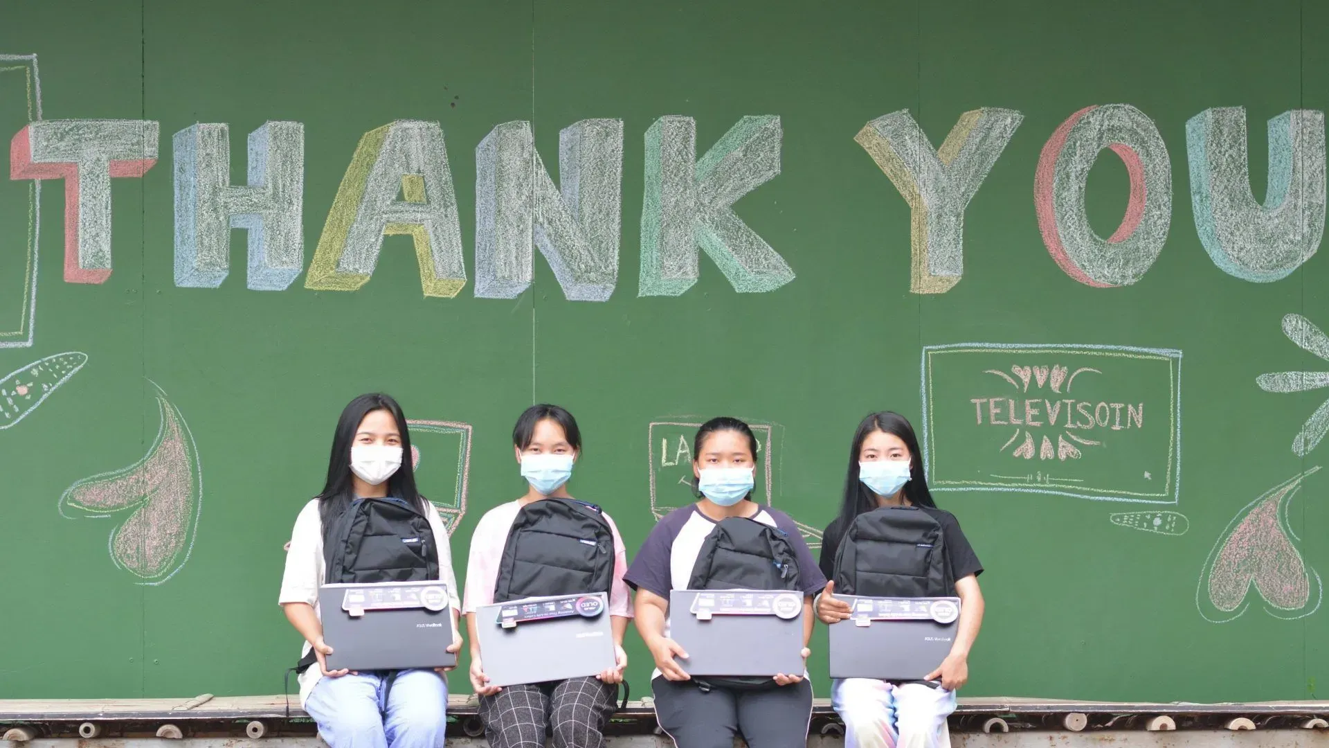 A group of young women wearing face masks are holding laptops in front of a chalkboard that says `` thank you ''.