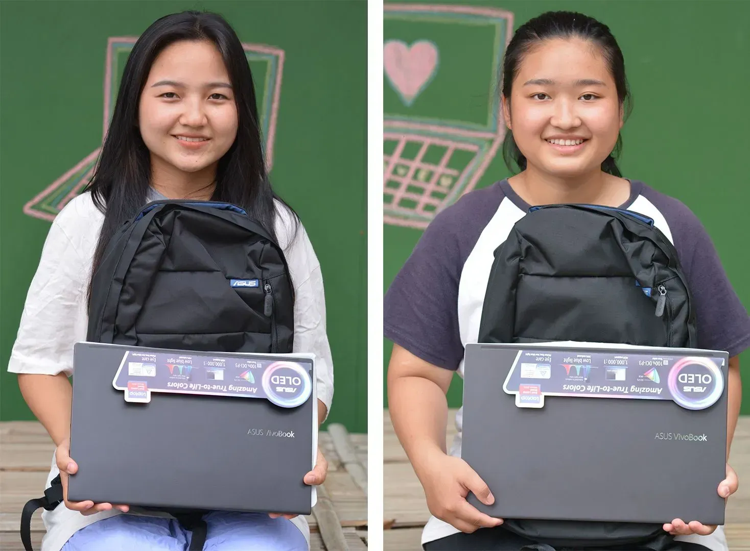 Two girls with backpacks are holding laptops in front of a green wall.