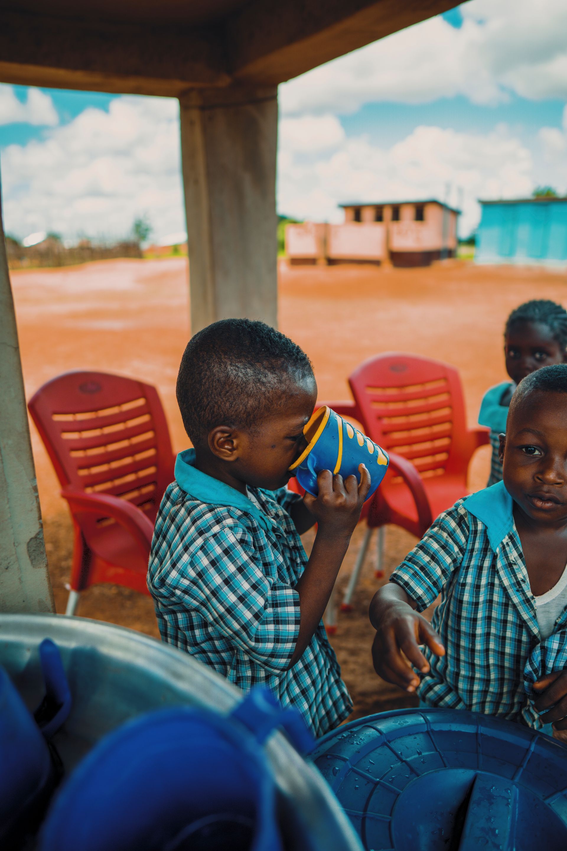Children drinking from blue cups outdoors near red chairs.