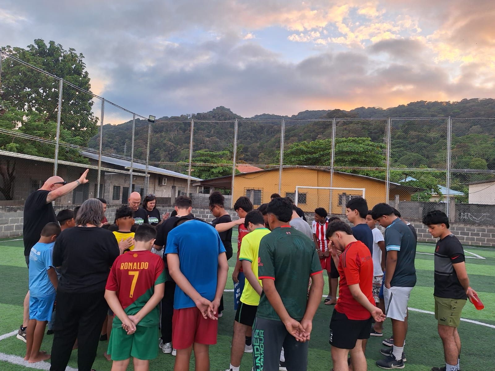 Group of people gathered on a soccer field at sunset, facing a coach near the goal.