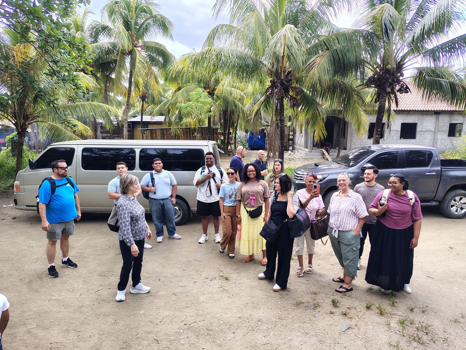 Group of people gathered outdoors beside white vans and a black SUV under palm trees.