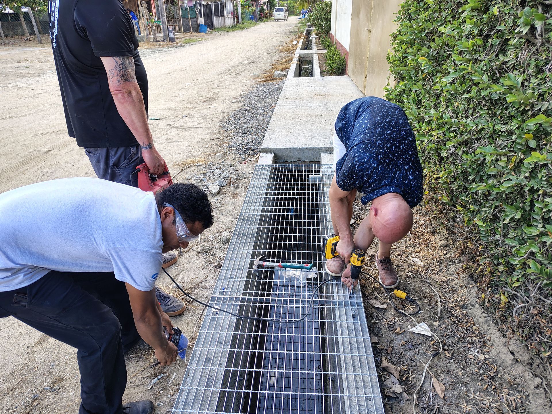 Three workers installing metal grates in a roadside drainage trench outdoors