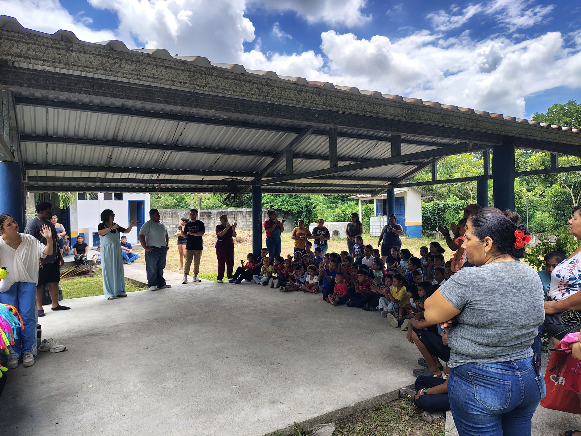 Crowd gathered under a metal-roofed pavilion, with people seated on one side and standing on the other.