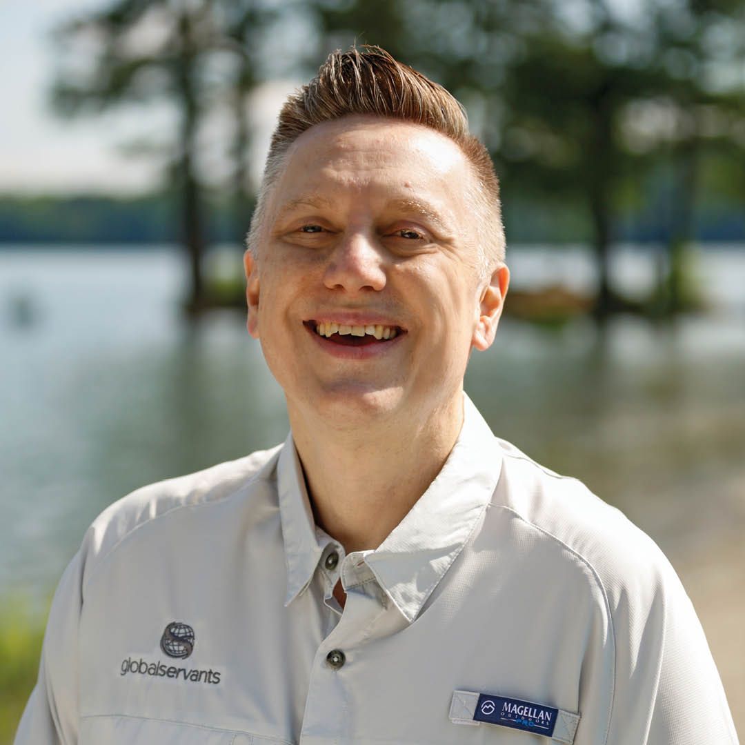 Travis Rutland smiling broadly in a light gray shirt, standing near a lake.