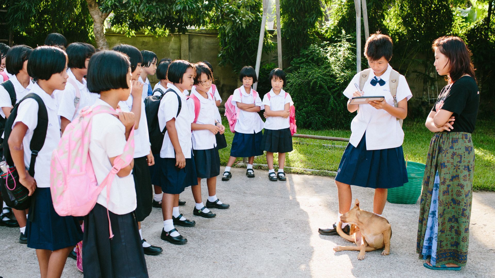 Students in school uniforms gather, a student holds a tablet, a teacher watches, and a dog rests on the ground.