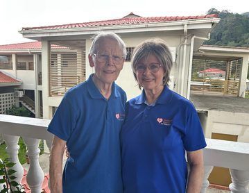 Two people in blue polo shirts standing on a balcony, smiling in front of a building with a red roof.