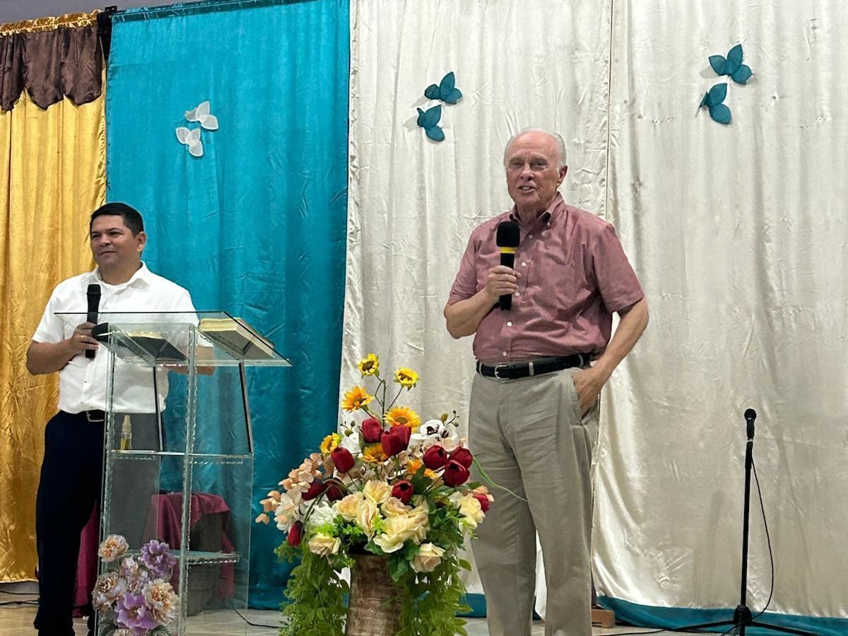 Two men speaking on stage beside a flower arrangement and blue-white draped backdrop