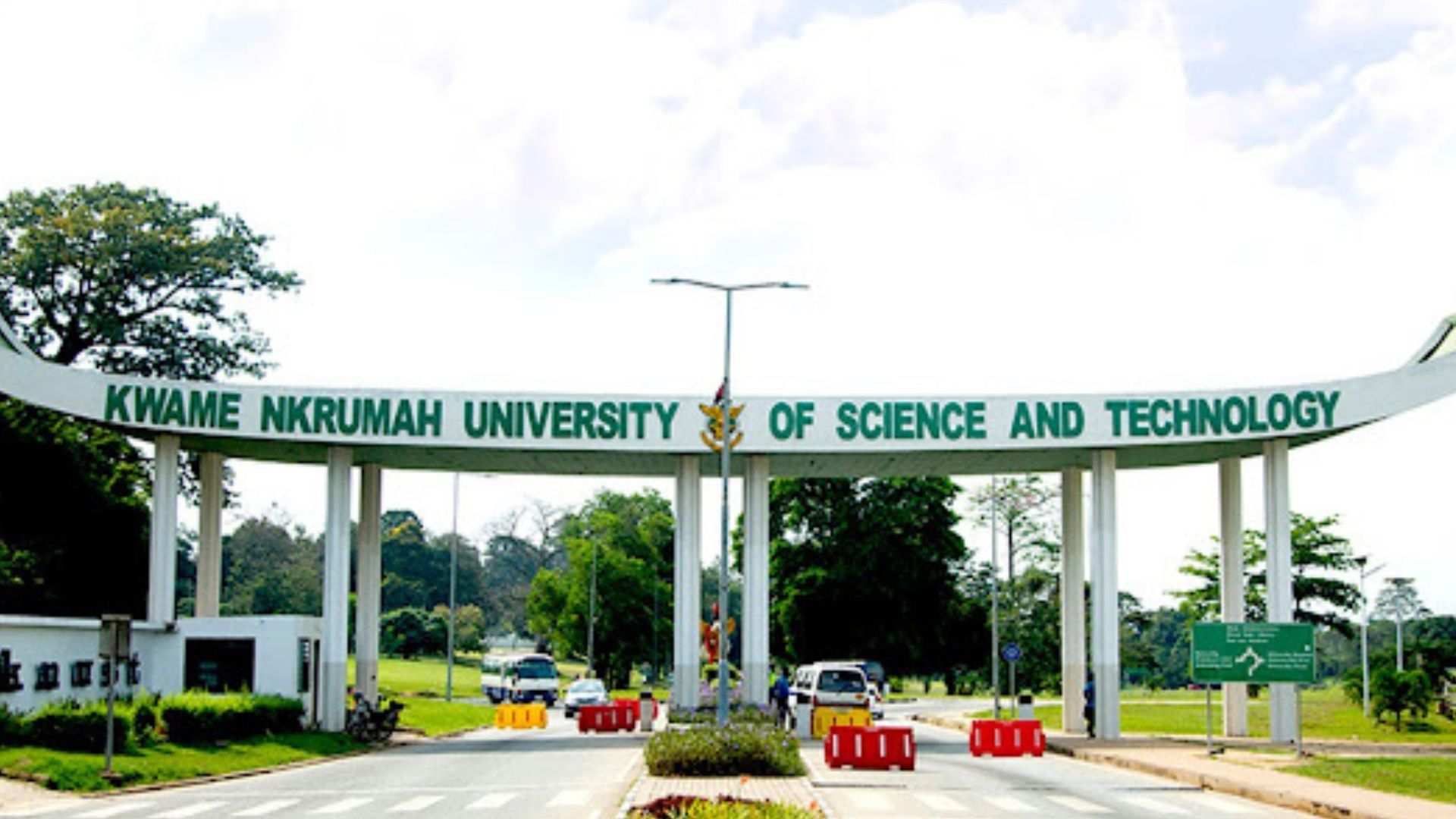 Entrance gate of Kwame Nkrumah University of Science and Technology. Green archway with text, road, and trees.