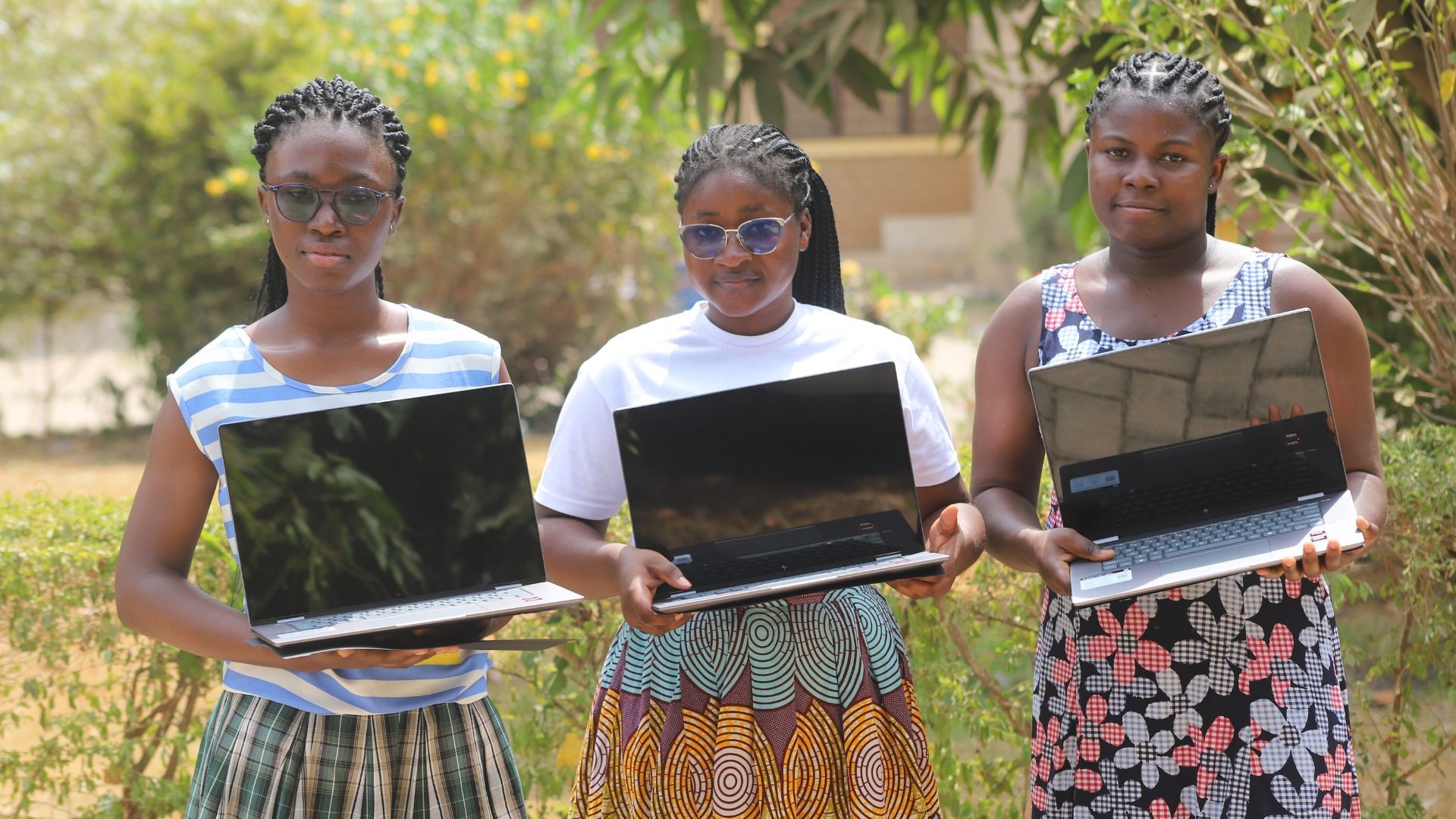 Three people holding up laptops outside in sunlight.