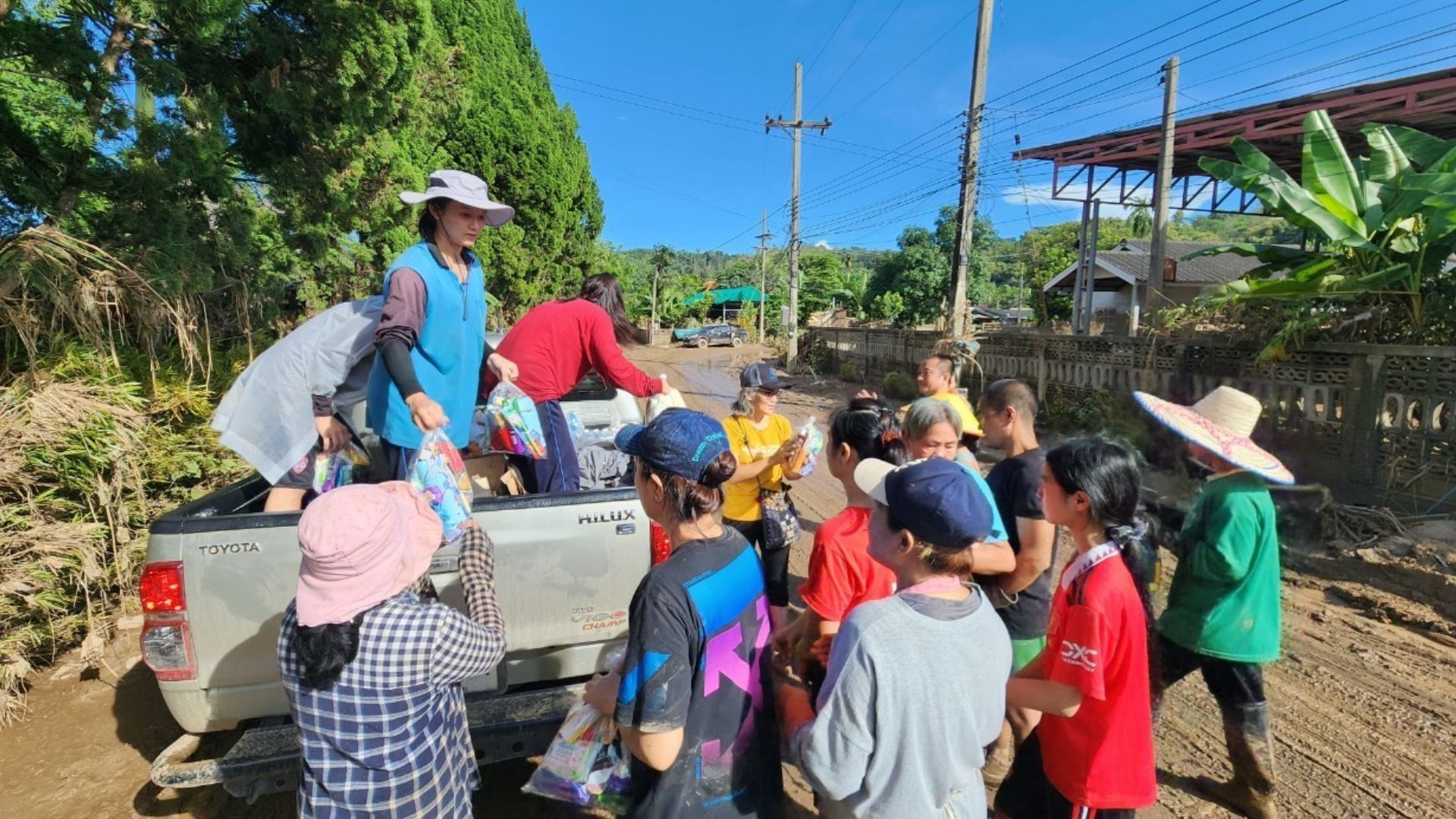 People distributing supplies from a truck in a flooded, muddy area with a building and greenery in the background.