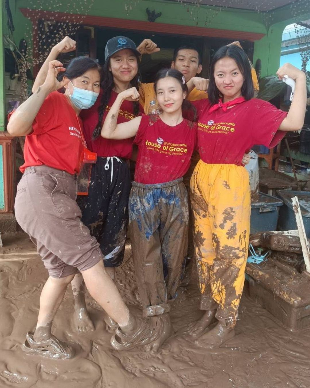 People in red shirts flexing in mud, smiling. Buildings in the background.